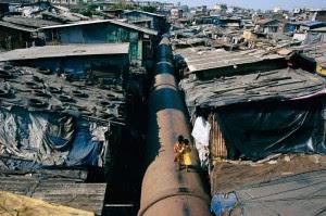 Photo with a immense pipe running through the middle surrounded by a shantytown with a woman and two children walking above the pipe.